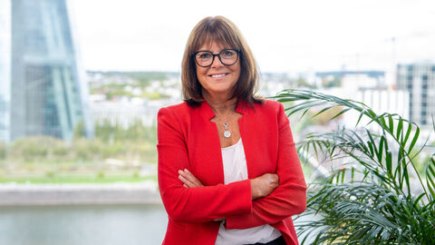 Eine lächelnde Frau mit mittellangen Haaren und Brille steht selbstbewusst mit verschränkten Armen in einem Büroumfeld. Sie trägt eine rote Blazerjacke über einem weißen Oberteil und eine Halskette. 