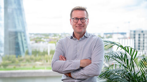 Ein lächelnder Mann mit kurzen Haaren und Brille steht selbstbewusst mit verschränkten Armen in einem Büroumfeld. Er trägt ein helles, gemustertes Hemd.